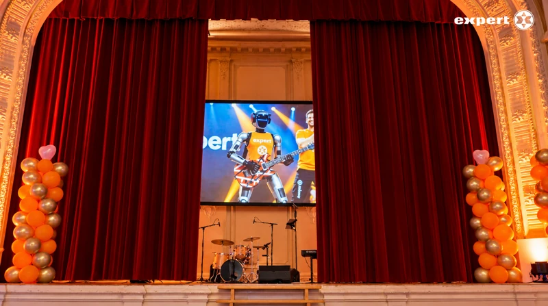 Galabühne mit rotem Vorhang, orange-weißen Ballons und digitalem Robotergitarristen auf der Leinwand
