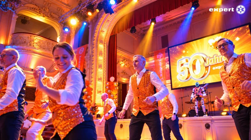 expert-Tanzteam in orange-goldenen Westen tanzt vor der Jubiläumsleinwand