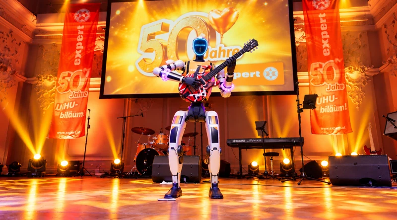 Unitree G1 mit E-Gitarre mittig auf der Bühne vor der goldenen 50-Jahre-expert-Projektion