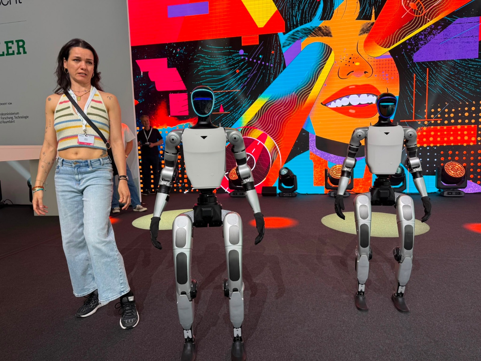 Zwei Unitree G1 Roboter mit junger Besucherin vor farbiger Bühne